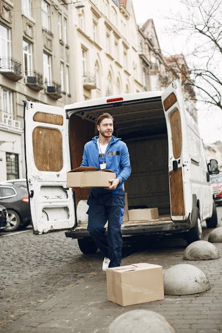 A man dressed in a blue jacket and dark trousers is standing outside a white moving van with its rear doors open on a cobblestone street in an urban area lined with multi-storey residential buildings. He is holding a cardboard box, preparing to load or unload it from the van. Additional cardboard boxes and packing materials are placed on the ground nearby, along with protective covering blankets draped inside the van. The scene captures the process of home relocation or furniture transport as part of a house removal service. The environment suggests daytime with natural light illuminating the scene, and the presence of parked cars and urban infrastructure indicating a residential area suitable for moving and packing services at [COMPANY_NAME].