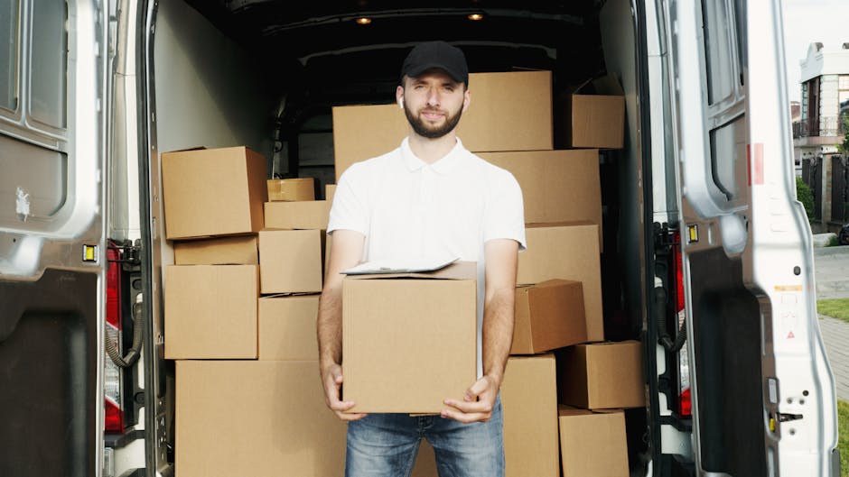 A man wearing a white shirt, dark cap, and jeans stands in the open back of a large white moving van, holding a cardboard box in front of him. Inside the van, multiple cardboard boxes of various sizes are stacked on the floor and along the sides, some covered with packing paper or protective materials. The scene captures part of the interior of the van with a clear view of the boxes being prepared for home relocation, with additional boxes visible towards the rear. Outside, the setting shows an outdoor pavement area with residential buildings partially visible in the background. The lighting indicates natural daylight, typical for a moving or packing process, demonstrating the logistics involved in furniture transport and packing during house removals, as handled by Man and Van Dartford.
