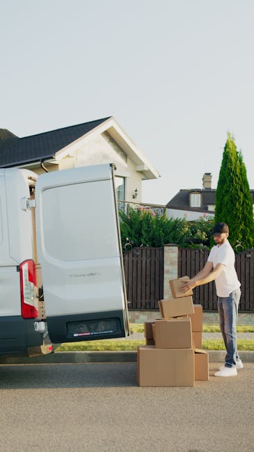 A man dressed in a blue jacket and dark trousers is standing outside a white moving van with its rear doors open on a cobblestone street in an urban area lined with multi-storey residential buildings. He is holding a cardboard box, preparing to load or unload it from the van. Additional cardboard boxes and packing materials are placed on the ground nearby, along with protective covering blankets draped inside the van. The scene captures the process of home relocation or furniture transport as part of a house removal service. The environment suggests daytime with natural light illuminating the scene, and the presence of parked cars and urban infrastructure indicating a residential area suitable for moving and packing services at [COMPANY_NAME].