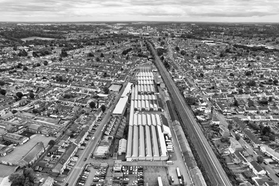 A black-and-white aerial photograph of Dartford railway station and surrounding residential area. The station features long, parallel train platforms with metal roofing, adjacent to a railway line that runs through the image. Nearby, various buildings including houses, shops, and industrial structures are visible, with narrow streets and parked cars in between. The surrounding neighborhood consists of tightly packed terraced houses with small front gardens, trees, and paved roads. In the background, open green spaces and distant landscape extend towards the horizon under a cloudy sky. This view captures the urban environment crucial for house removals and efficient furniture transport, illustrating the area's infrastructure that Man and Van Dartford uses for home relocation and packing services.