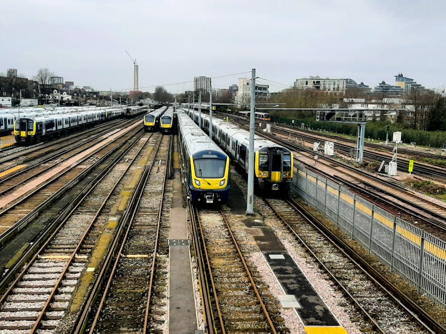 A black-and-white aerial photograph of Dartford railway station and surrounding residential area. The station features long, parallel train platforms with metal roofing, adjacent to a railway line that runs through the image. Nearby, various buildings including houses, shops, and industrial structures are visible, with narrow streets and parked cars in between. The surrounding neighborhood consists of tightly packed terraced houses with small front gardens, trees, and paved roads. In the background, open green spaces and distant landscape extend towards the horizon under a cloudy sky. This view captures the urban environment crucial for house removals and efficient furniture transport, illustrating the area's infrastructure that Man and Van Dartford uses for home relocation and packing services.